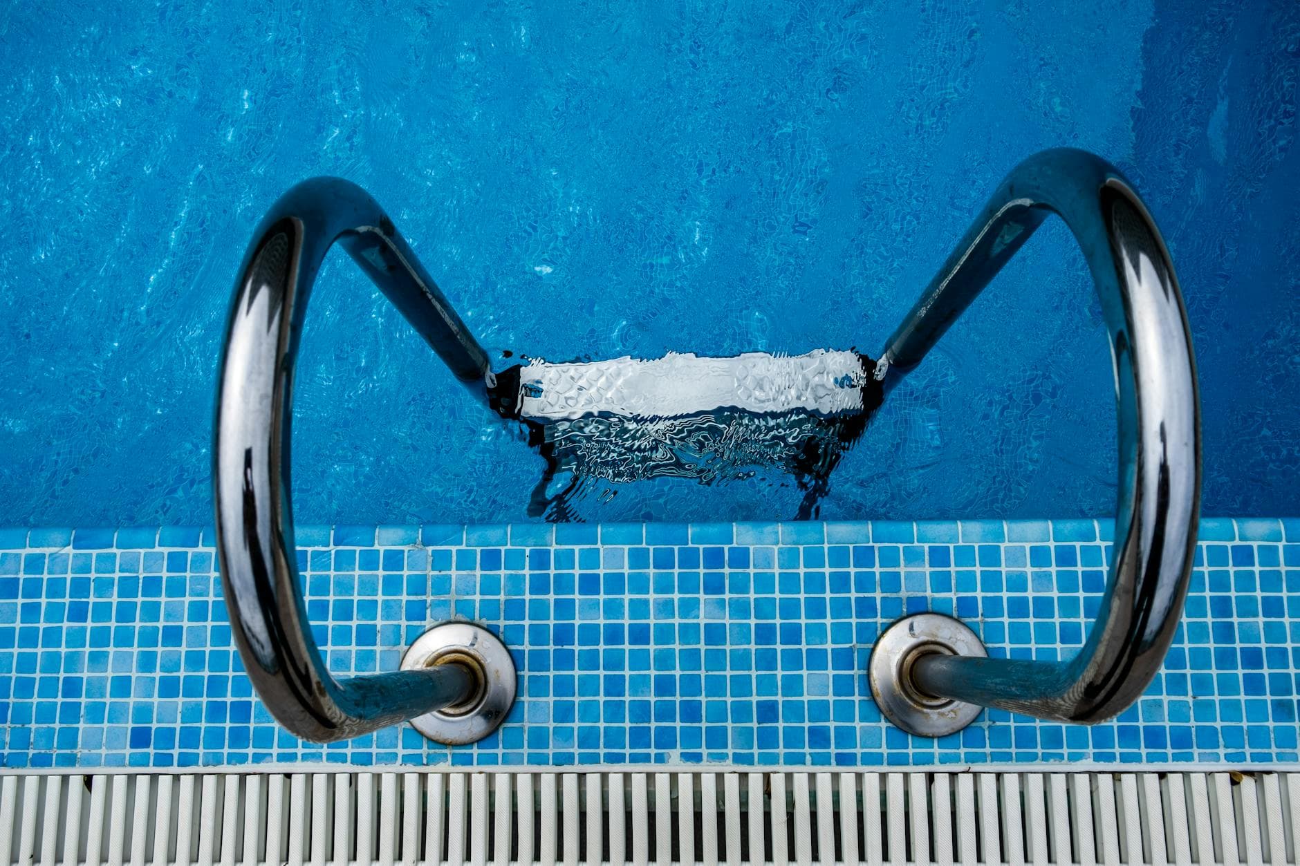 Aerial view of a stainless steel ladder entering a sparkling blue swimming pool with tiled edges.