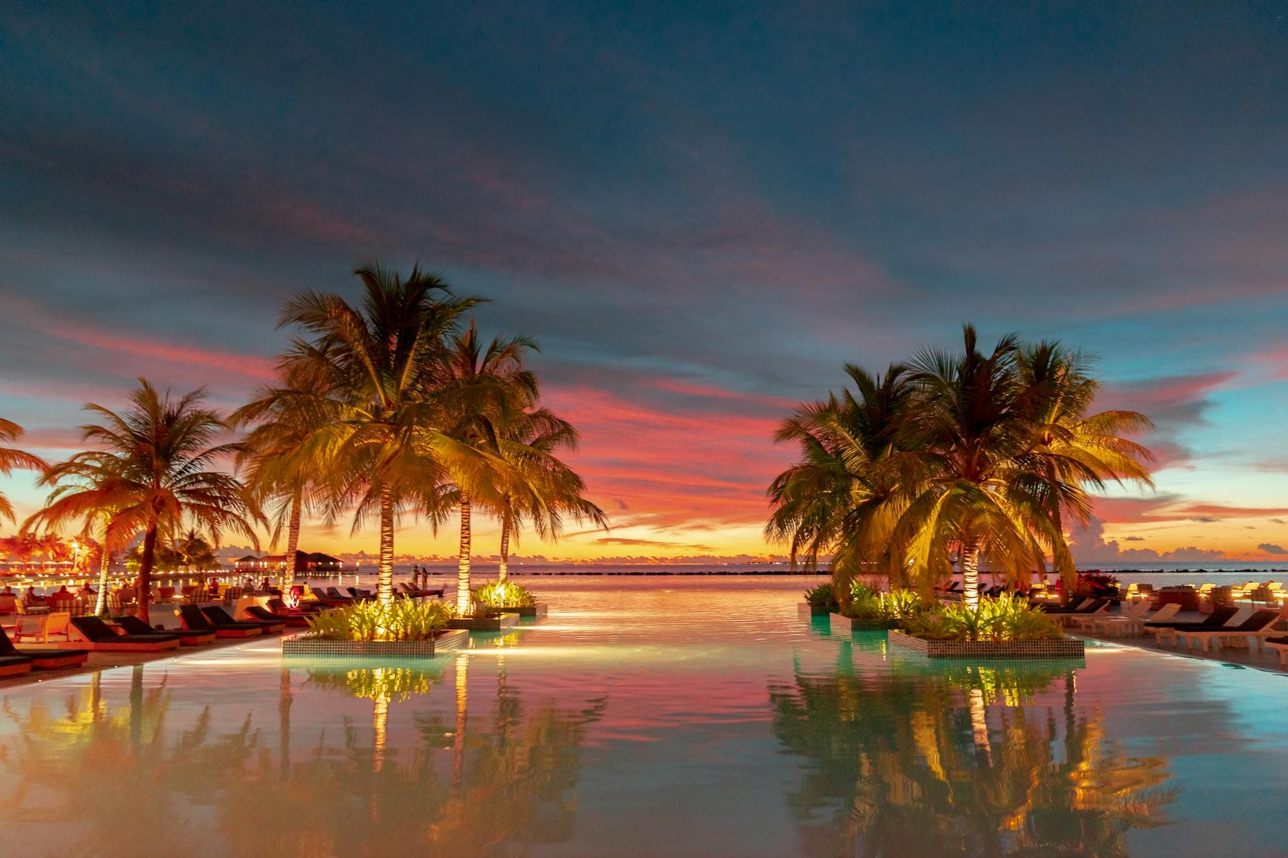 Beautiful sunset over a tropical resort pool with palm trees and vibrant sky reflections.